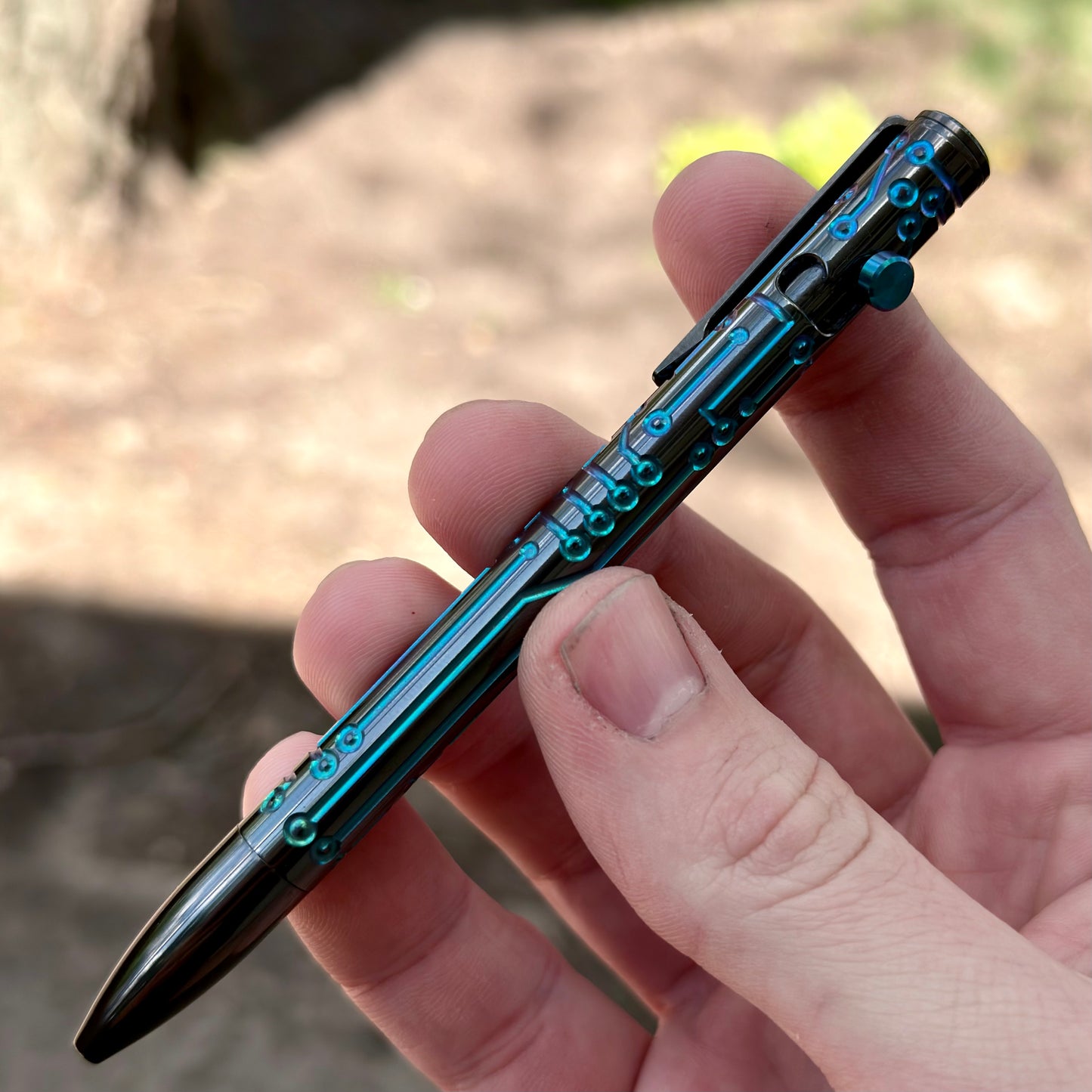 A close-up of a hand holding a metallic ballpoint pen with a dark teal and black cybernetic or circuit-like pattern, set against a blurred outdoor background.