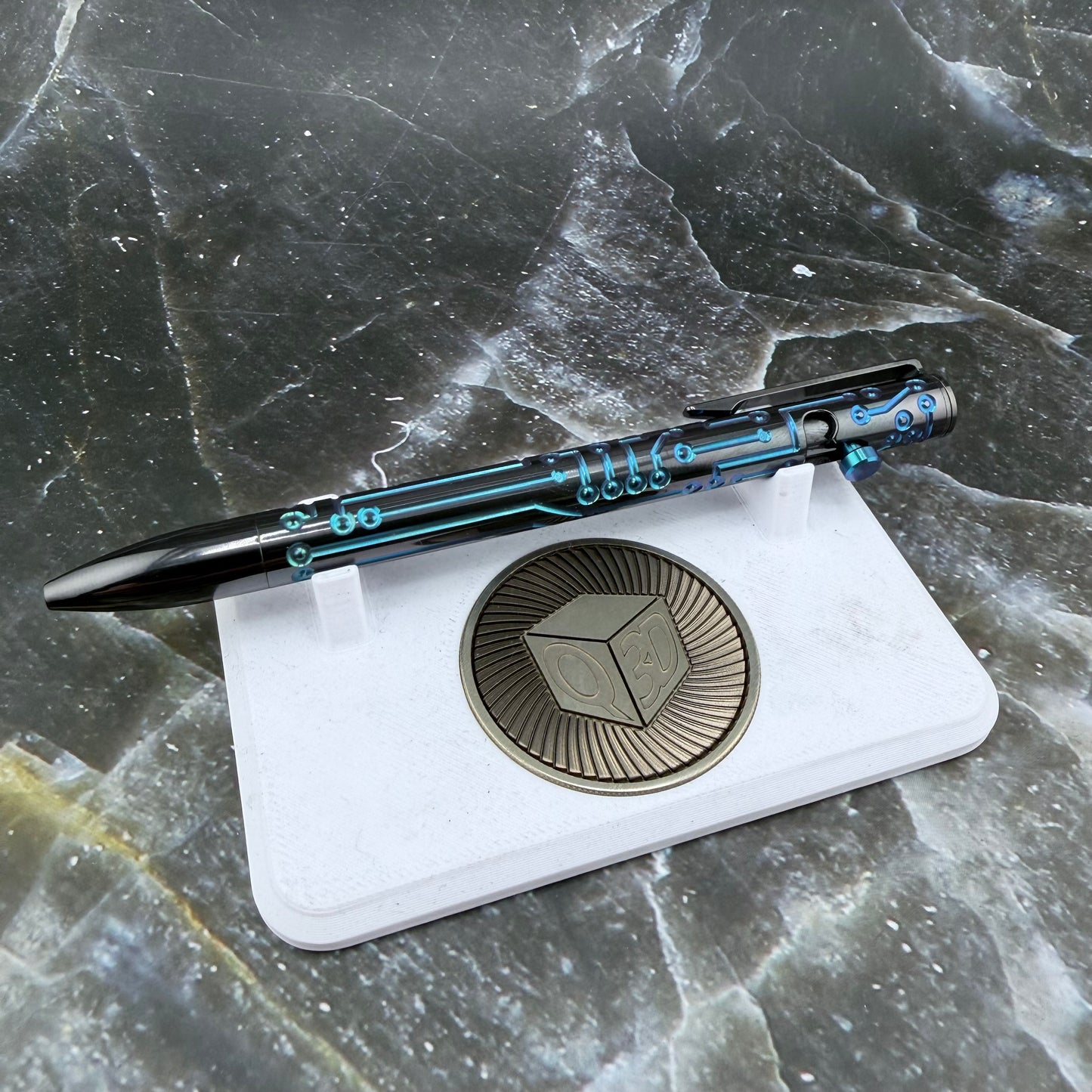 A black pen with blue circuit-like graphics rests on a white display stand featuring a circular metallic emblem, set against a dark marbled surface.