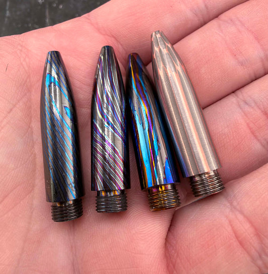 Four metallic bullet-shapedpen tips displayed on an open hand. Three have iridescent blue, purple, and silver striped finishes, and one is a brushed silver metal.