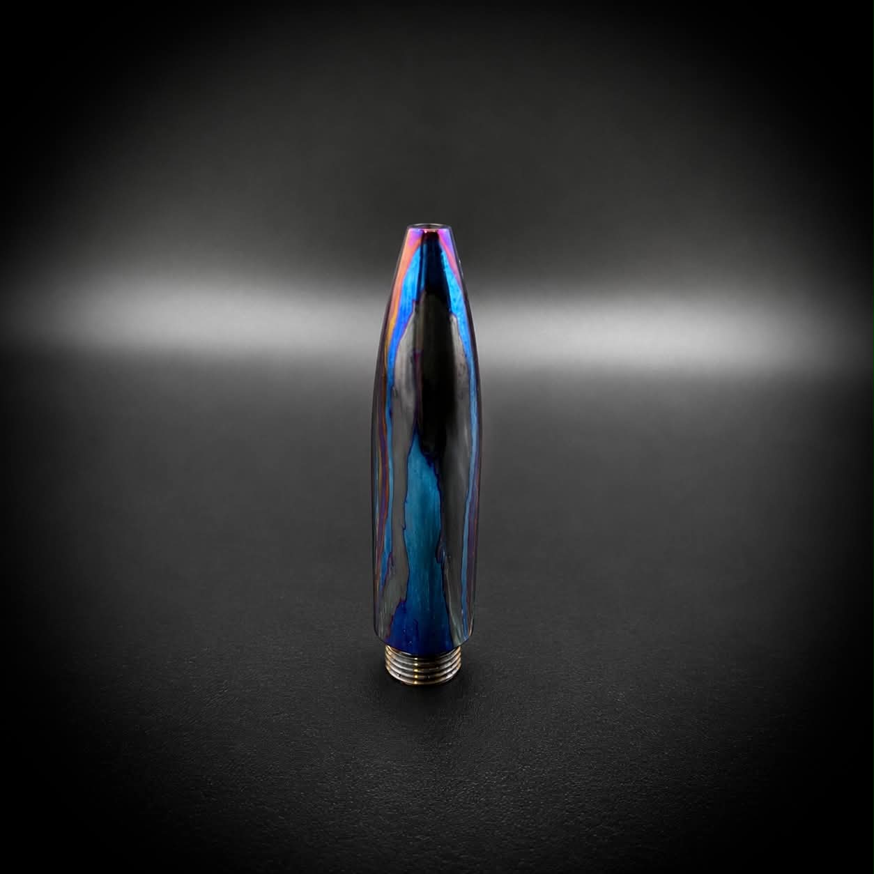 A metallic bullet-shaped pen tip with a dark blue and black base and an iridescent, rainbow-reflective coating, standing upright on a dark, vignetted background.