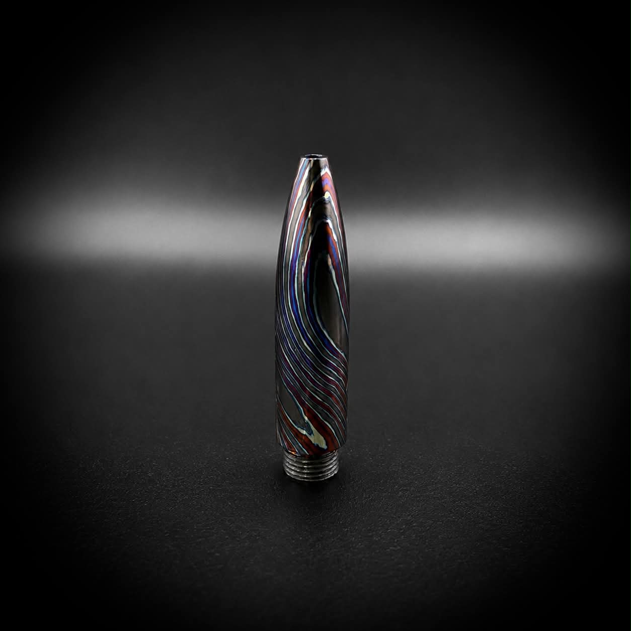 A single bullet-shaped pen tip with a metallic silver base, featuring a black and iridescent multicolor swirl pattern, set against a dark studio background with a spotlight effect.