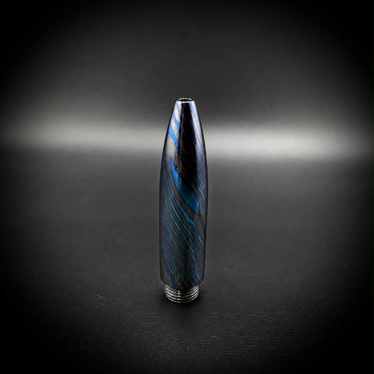 A metallic bullet-shaped pen tip with a blue and black marbled pattern, standing upright on a dark surface against a dark background.
